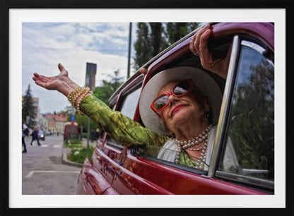 A stylish elderly woman wearing a white hat, red sunglasses, and pearl necklaces leans out of the passenger window of a classic red car. Her arm is outstretched with a joyful expression on her face, enjoying a ride through a city street. Poster