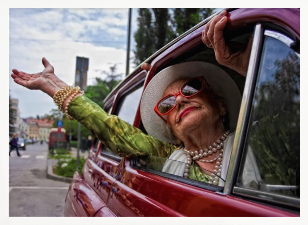A stylish elderly woman wearing a white hat, red sunglasses, and pearl necklaces leans out of the passenger window of a classic red car. Her arm is outstretched with a joyful expression on her face, enjoying a ride through a city street. Poster
