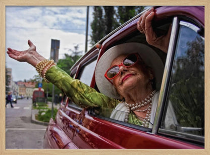 A stylish elderly woman wearing a white hat, red sunglasses, and pearl necklaces leans out of the passenger window of a classic red car. Her arm is outstretched with a joyful expression on her face, enjoying a ride through a city street. Poster