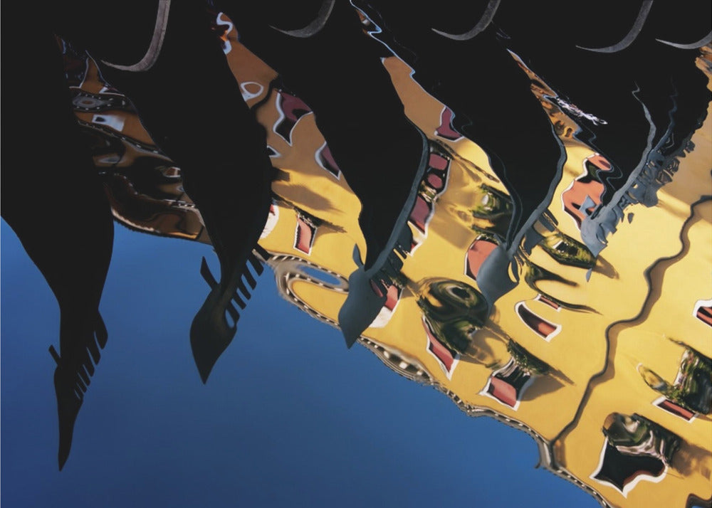 An abstract photograph of a distorted reflection in water, showing a vibrant yellow building against a deep blue sky, with the dark silhouettes of several Venetian gondola prows cutting across the top of the image. The entire scene is enclosed in a silver picture frame. Print