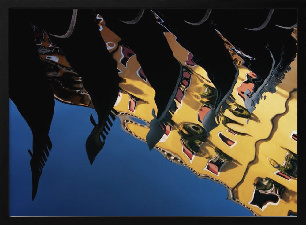 An abstract photograph of a distorted reflection in water, showing a vibrant yellow building against a deep blue sky, with the dark silhouettes of several Venetian gondola prows cutting across the top of the image. The entire scene is enclosed in a silver picture frame. Print