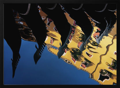 An abstract photograph of a distorted reflection in water, showing a vibrant yellow building against a deep blue sky, with the dark silhouettes of several Venetian gondola prows cutting across the top of the image. The entire scene is enclosed in a silver picture frame. Print