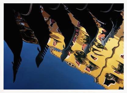 An abstract photograph of a distorted reflection in water, showing a vibrant yellow building against a deep blue sky, with the dark silhouettes of several Venetian gondola prows cutting across the top of the image. The entire scene is enclosed in a silver picture frame. Print