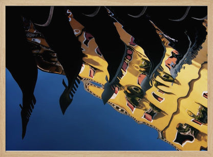 An abstract photograph of a distorted reflection in water, showing a vibrant yellow building against a deep blue sky, with the dark silhouettes of several Venetian gondola prows cutting across the top of the image. The entire scene is enclosed in a silver picture frame. Print