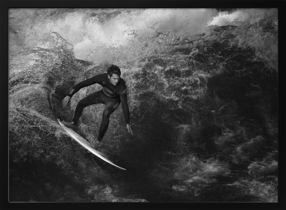 A dramatic black and white photograph of a surfer in a wetsuit skillfully riding down the face of a large, turbulent wave. The image is enclosed in a silver frame. Wall Art