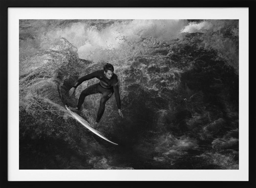 A dramatic black and white photograph of a surfer in a wetsuit skillfully riding down the face of a large, turbulent wave. The image is enclosed in a silver frame. Wall Art