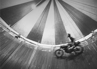 A dynamic, wide-angle black and white photograph of a motorcycle rider performing a stunt in a motordrome, commonly known as the 'wall of death'. The rider is a blur of motion as they speed along the steep, curved wooden walls. The perspective looks up towards the striped canvas top of the tent, with a few spectators visible watching from the railing at the top edge. The entire image is enclosed in a silver frame. Print