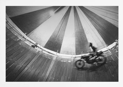 A dynamic, wide-angle black and white photograph of a motorcycle rider performing a stunt in a motordrome, commonly known as the 'wall of death'. The rider is a blur of motion as they speed along the steep, curved wooden walls. The perspective looks up towards the striped canvas top of the tent, with a few spectators visible watching from the railing at the top edge. The entire image is enclosed in a silver frame. Print