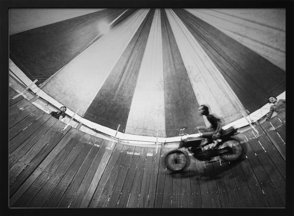A dynamic, wide-angle black and white photograph of a motorcycle rider performing a stunt in a motordrome, commonly known as the 'wall of death'. The rider is a blur of motion as they speed along the steep, curved wooden walls. The perspective looks up towards the striped canvas top of the tent, with a few spectators visible watching from the railing at the top edge. The entire image is enclosed in a silver frame. Print