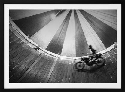 A dynamic, wide-angle black and white photograph of a motorcycle rider performing a stunt in a motordrome, commonly known as the 'wall of death'. The rider is a blur of motion as they speed along the steep, curved wooden walls. The perspective looks up towards the striped canvas top of the tent, with a few spectators visible watching from the railing at the top edge. The entire image is enclosed in a silver frame. Print