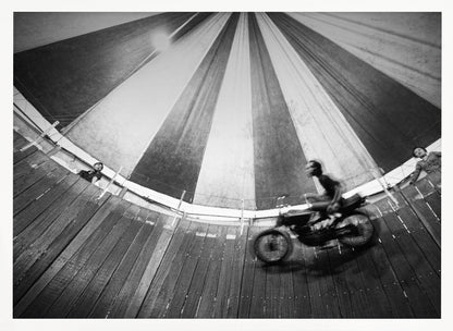 A dynamic, wide-angle black and white photograph of a motorcycle rider performing a stunt in a motordrome, commonly known as the 'wall of death'. The rider is a blur of motion as they speed along the steep, curved wooden walls. The perspective looks up towards the striped canvas top of the tent, with a few spectators visible watching from the railing at the top edge. The entire image is enclosed in a silver frame. Print