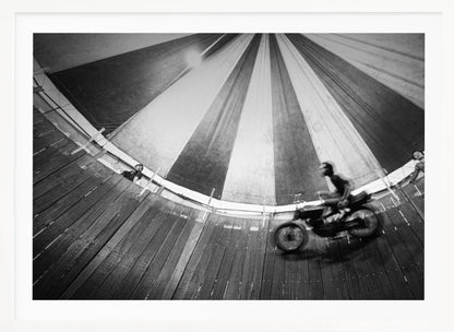 A dynamic, wide-angle black and white photograph of a motorcycle rider performing a stunt in a motordrome, commonly known as the 'wall of death'. The rider is a blur of motion as they speed along the steep, curved wooden walls. The perspective looks up towards the striped canvas top of the tent, with a few spectators visible watching from the railing at the top edge. The entire image is enclosed in a silver frame. Print