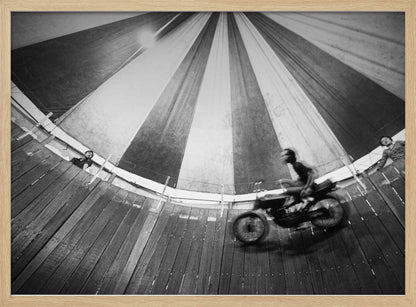 A dynamic, wide-angle black and white photograph of a motorcycle rider performing a stunt in a motordrome, commonly known as the 'wall of death'. The rider is a blur of motion as they speed along the steep, curved wooden walls. The perspective looks up towards the striped canvas top of the tent, with a few spectators visible watching from the railing at the top edge. The entire image is enclosed in a silver frame. Print