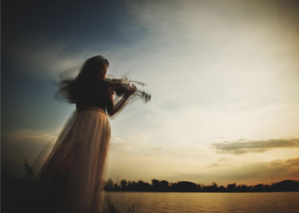 An artistic, framed photograph with motion blur capturing the silhouette of a woman in a long flowing skirt playing the violin by a lake against a dramatic sunset sky. Poster