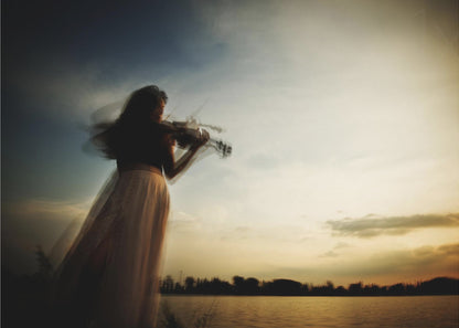 An artistic, framed photograph with motion blur capturing the silhouette of a woman in a long flowing skirt playing the violin by a lake against a dramatic sunset sky. Poster