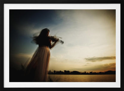 An artistic, framed photograph with motion blur capturing the silhouette of a woman in a long flowing skirt playing the violin by a lake against a dramatic sunset sky. Poster
