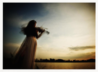 An artistic, framed photograph with motion blur capturing the silhouette of a woman in a long flowing skirt playing the violin by a lake against a dramatic sunset sky. Poster