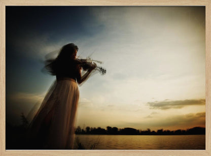 An artistic, framed photograph with motion blur capturing the silhouette of a woman in a long flowing skirt playing the violin by a lake against a dramatic sunset sky. Poster