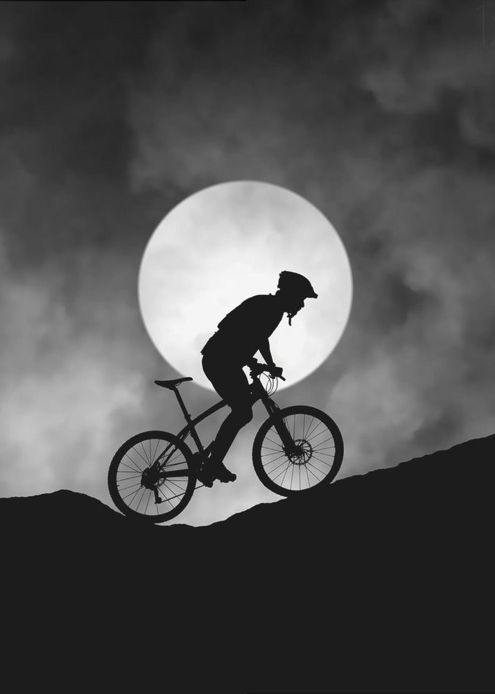 A dramatic black and white silhouette of a mountain biker riding uphill against the backdrop of a large, full moon in a cloudy night sky. Decor