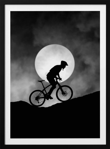 A dramatic black and white silhouette of a mountain biker riding uphill against the backdrop of a large, full moon in a cloudy night sky. Decor