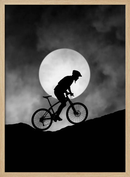 A dramatic black and white silhouette of a mountain biker riding uphill against the backdrop of a large, full moon in a cloudy night sky. Decor