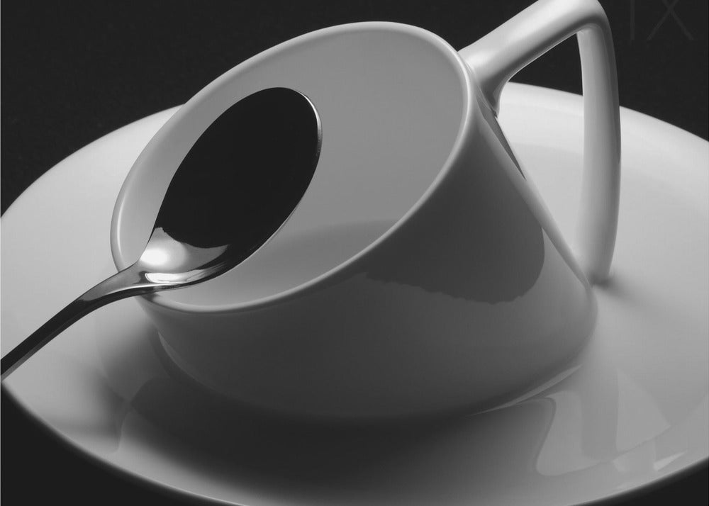A dramatic black and white close-up photograph of a white porcelain coffee cup tilted on its saucer, with a reflective spoon resting inside. The image has high contrast against a dark background and is presented within a silver-colored frame. Decor
