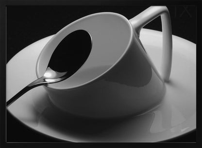 A dramatic black and white close-up photograph of a white porcelain coffee cup tilted on its saucer, with a reflective spoon resting inside. The image has high contrast against a dark background and is presented within a silver-colored frame. Decor