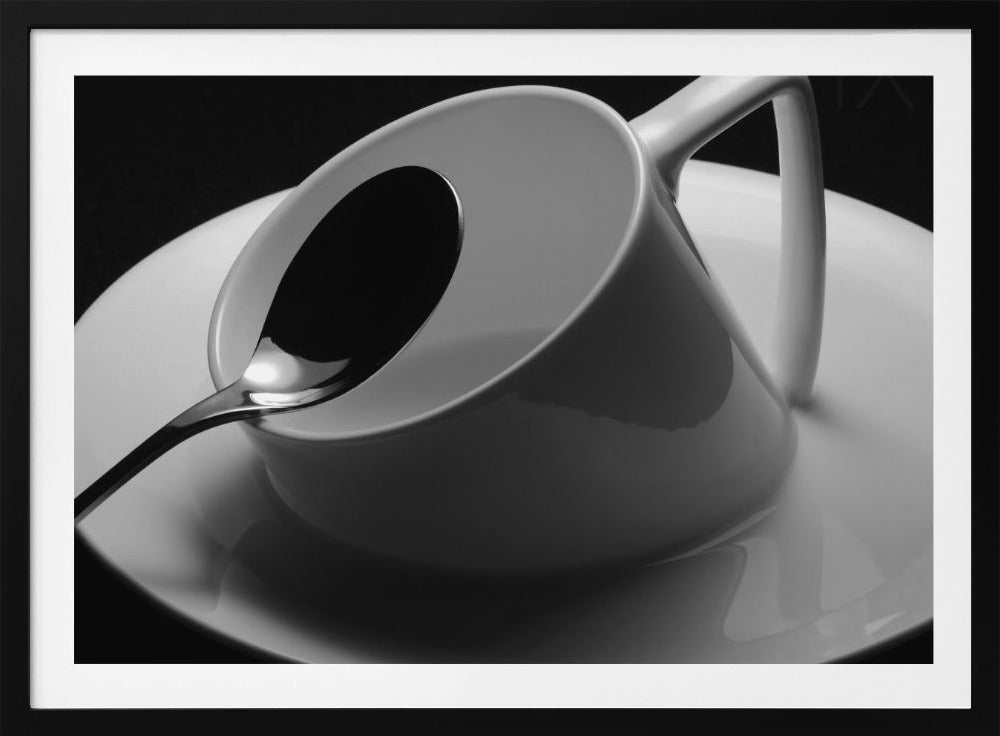 A dramatic black and white close-up photograph of a white porcelain coffee cup tilted on its saucer, with a reflective spoon resting inside. The image has high contrast against a dark background and is presented within a silver-colored frame. Decor