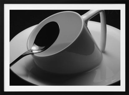 A dramatic black and white close-up photograph of a white porcelain coffee cup tilted on its saucer, with a reflective spoon resting inside. The image has high contrast against a dark background and is presented within a silver-colored frame. Decor