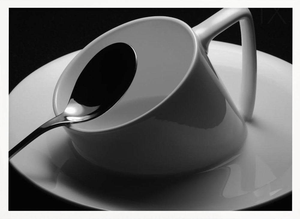 A dramatic black and white close-up photograph of a white porcelain coffee cup tilted on its saucer, with a reflective spoon resting inside. The image has high contrast against a dark background and is presented within a silver-colored frame. Decor
