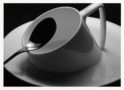 A dramatic black and white close-up photograph of a white porcelain coffee cup tilted on its saucer, with a reflective spoon resting inside. The image has high contrast against a dark background and is presented within a silver-colored frame. Decor