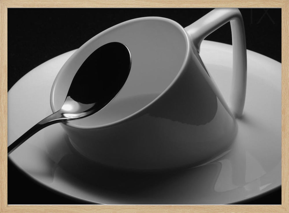 A dramatic black and white close-up photograph of a white porcelain coffee cup tilted on its saucer, with a reflective spoon resting inside. The image has high contrast against a dark background and is presented within a silver-colored frame. Decor