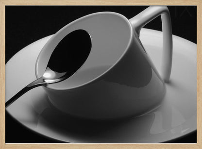 A dramatic black and white close-up photograph of a white porcelain coffee cup tilted on its saucer, with a reflective spoon resting inside. The image has high contrast against a dark background and is presented within a silver-colored frame. Decor