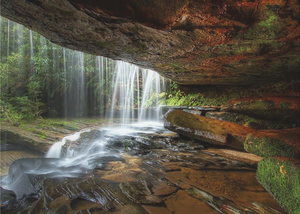 A long-exposure photograph taken from underneath a rock overhang, looking out through a cascading waterfall. The water appears silky as it flows over mossy rocks in a stream below. The scene is framed by a silver border. Print