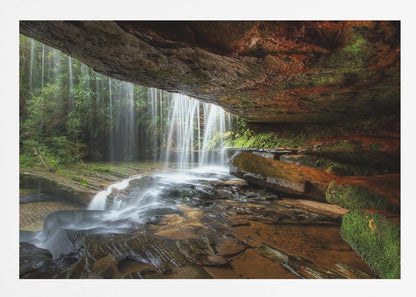 A long-exposure photograph taken from underneath a rock overhang, looking out through a cascading waterfall. The water appears silky as it flows over mossy rocks in a stream below. The scene is framed by a silver border. Print