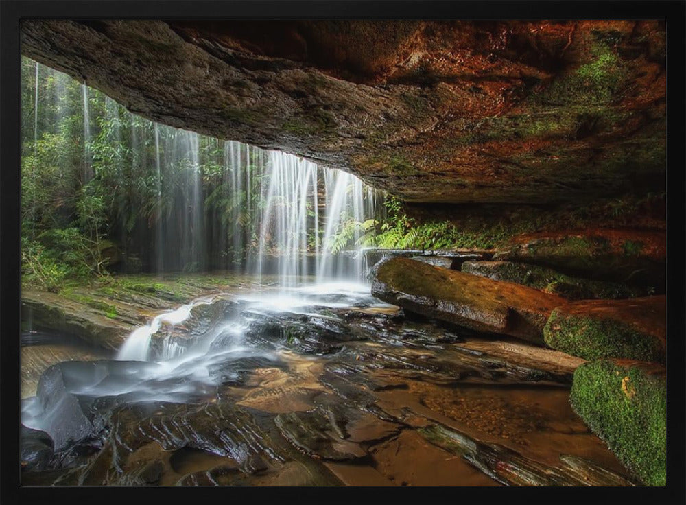 A long-exposure photograph taken from underneath a rock overhang, looking out through a cascading waterfall. The water appears silky as it flows over mossy rocks in a stream below. The scene is framed by a silver border. Print