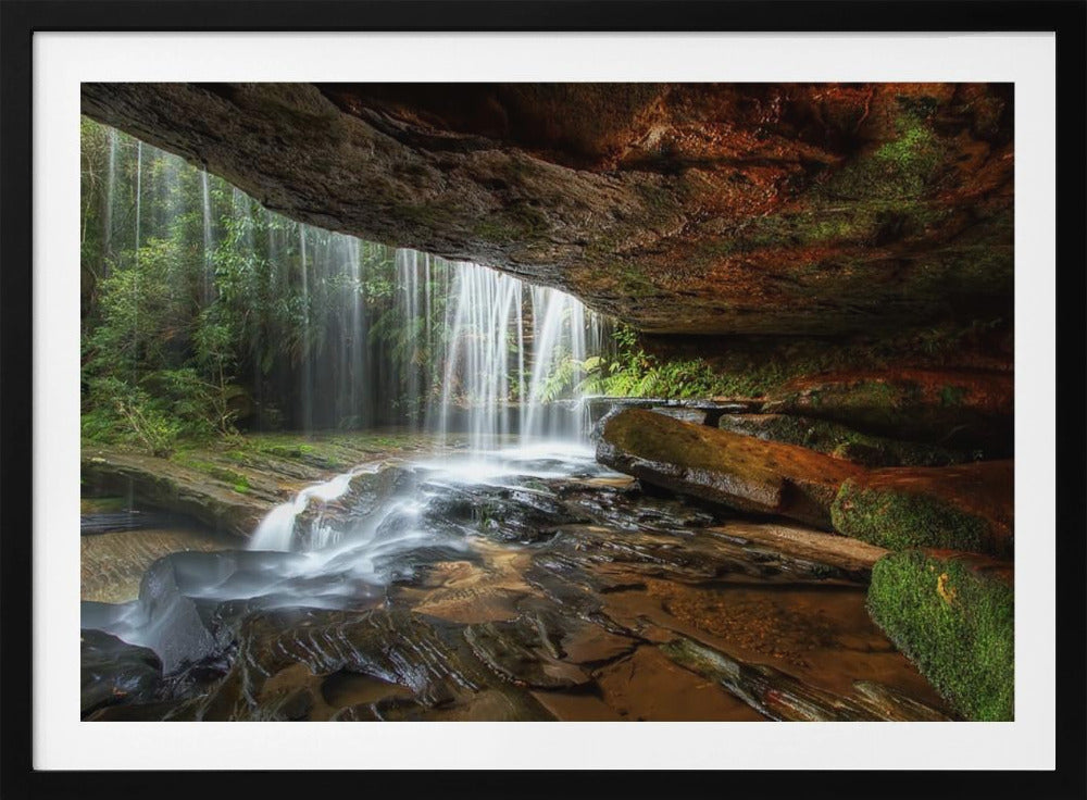 A long-exposure photograph taken from underneath a rock overhang, looking out through a cascading waterfall. The water appears silky as it flows over mossy rocks in a stream below. The scene is framed by a silver border. Print