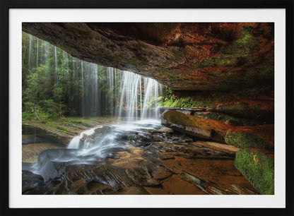 A long-exposure photograph taken from underneath a rock overhang, looking out through a cascading waterfall. The water appears silky as it flows over mossy rocks in a stream below. The scene is framed by a silver border. Print
