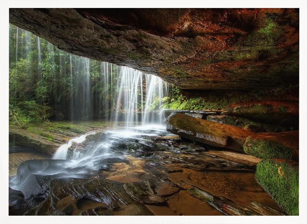 A long-exposure photograph taken from underneath a rock overhang, looking out through a cascading waterfall. The water appears silky as it flows over mossy rocks in a stream below. The scene is framed by a silver border. Print