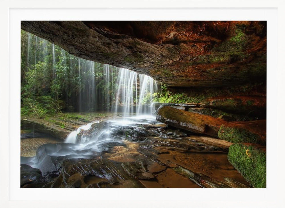 A long-exposure photograph taken from underneath a rock overhang, looking out through a cascading waterfall. The water appears silky as it flows over mossy rocks in a stream below. The scene is framed by a silver border. Print