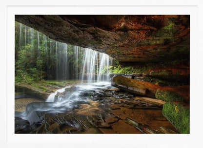 A long-exposure photograph taken from underneath a rock overhang, looking out through a cascading waterfall. The water appears silky as it flows over mossy rocks in a stream below. The scene is framed by a silver border. Print