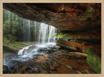 A long-exposure photograph taken from underneath a rock overhang, looking out through a cascading waterfall. The water appears silky as it flows over mossy rocks in a stream below. The scene is framed by a silver border. Print