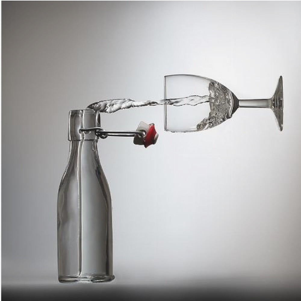 A conceptual photograph in a light wood frame depicting water pouring horizontally from an open swing-top glass bottle into a floating, sideways wine glass against a simple gray background, capturing the liquid in mid-air. Print
