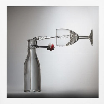 A conceptual photograph in a light wood frame depicting water pouring horizontally from an open swing-top glass bottle into a floating, sideways wine glass against a simple gray background, capturing the liquid in mid-air. Print