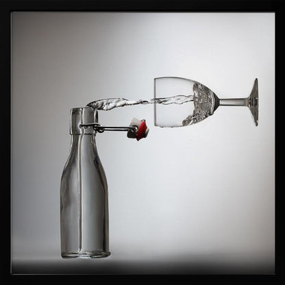 A conceptual photograph in a light wood frame depicting water pouring horizontally from an open swing-top glass bottle into a floating, sideways wine glass against a simple gray background, capturing the liquid in mid-air. Print