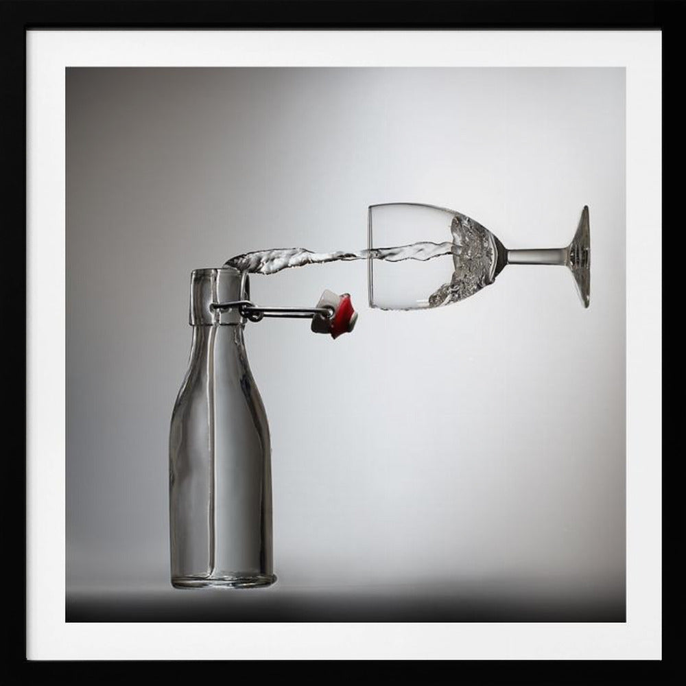 A conceptual photograph in a light wood frame depicting water pouring horizontally from an open swing-top glass bottle into a floating, sideways wine glass against a simple gray background, capturing the liquid in mid-air. Print