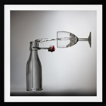 A conceptual photograph in a light wood frame depicting water pouring horizontally from an open swing-top glass bottle into a floating, sideways wine glass against a simple gray background, capturing the liquid in mid-air. Print