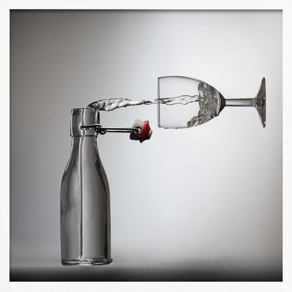A conceptual photograph in a light wood frame depicting water pouring horizontally from an open swing-top glass bottle into a floating, sideways wine glass against a simple gray background, capturing the liquid in mid-air. Print