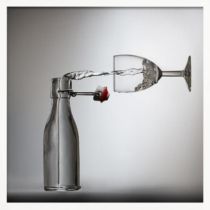 A conceptual photograph in a light wood frame depicting water pouring horizontally from an open swing-top glass bottle into a floating, sideways wine glass against a simple gray background, capturing the liquid in mid-air. Print