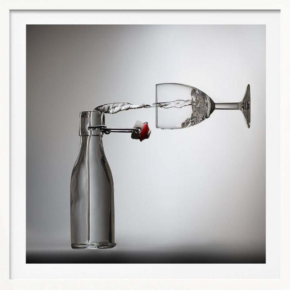 A conceptual photograph in a light wood frame depicting water pouring horizontally from an open swing-top glass bottle into a floating, sideways wine glass against a simple gray background, capturing the liquid in mid-air. Print